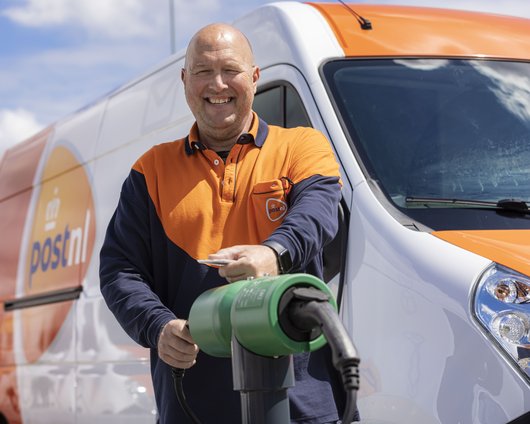 Smiling delivery driver in a uniform standing in front of an electric van, holding a charging cable and preparing to charge the vehicle.