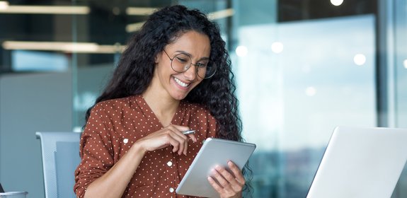 Smiling woman with curly hair and glasses working in a modern office, using a tablet and stylus while seated at a desk with a laptop and papers.