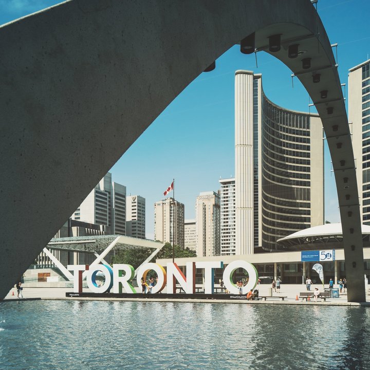 View of Toronto’s iconic city sign in front of Nathan Phillips Square reflecting pool, with modern curved towers of City Hall and a concrete arch framing the scene under a clear blue sky.